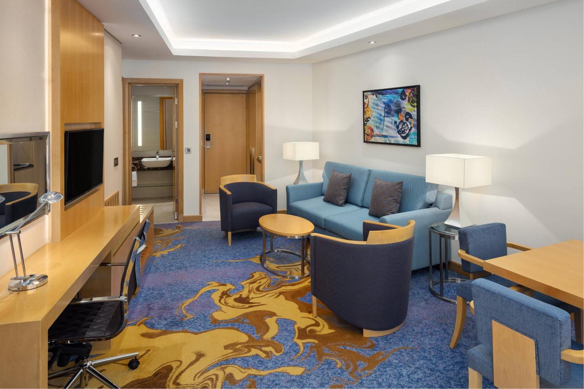 โรงแรมเชอราตัน มักกะฮ์ จาบาล อัล กาบา Mecca ภายนอก รูปภาพ The photo displays a modern hotel room or suite. The room features a comfortable seating area with a blue couch and two blue armchairs surrounding a small round table. A wooden desk with a black office chair is positioned against one wall, accompanie