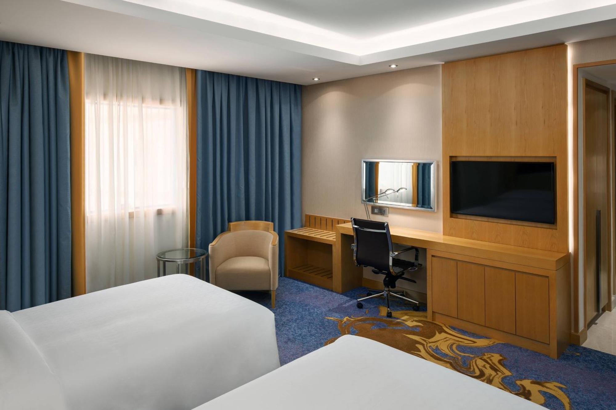 โรงแรมเชอราตัน มักกะฮ์ จาบาล อัล กาบา Mecca ภายนอก รูปภาพ The photo shows a hotel room featuring two beds with white linens. There are large windows covered with blue curtains that allow natural light to enter. On one side of the room, there is a wooden workstation with a chair and a mirror, along with a fl