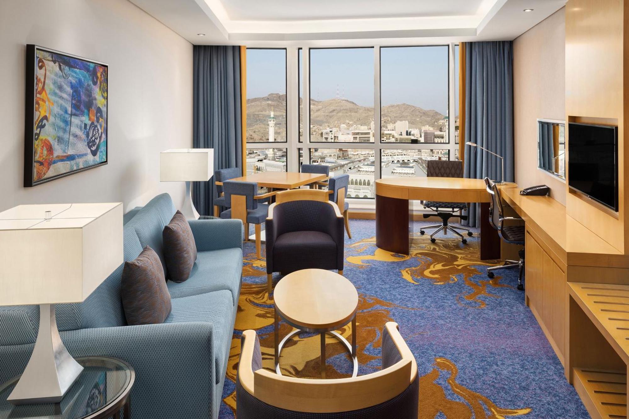 โรงแรมเชอราตัน มักกะฮ์ จาบาล อัล กาบา Mecca ภายนอก รูปภาพ The photo shows a well-decorated hotel room featuring a modern and comfortable interior. There is a cozy seating area with a blue sofa and armchairs, complemented by a round coffee table. A desk with a chair is also present, ideal for work. The room 