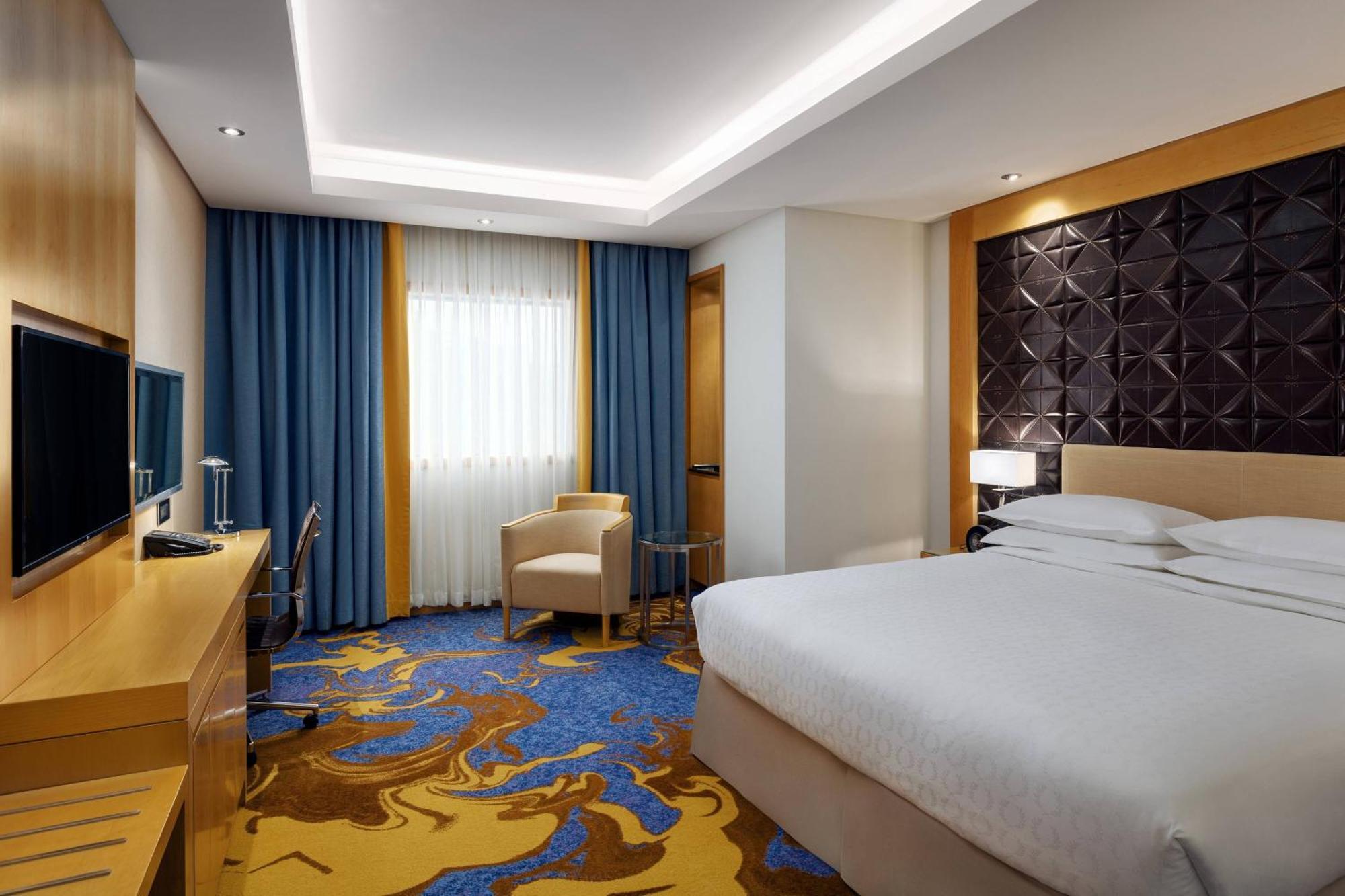 โรงแรมเชอราตัน มักกะฮ์ จาบาล อัล กาบา Mecca ภายนอก รูปภาพ The photo shows a modern hotel room featuring a comfortable double bed with a light-colored headboard. There's a large window dressed with blue curtains, allowing natural light to enter. The walls are painted in neutral tones, and one wall has a text
