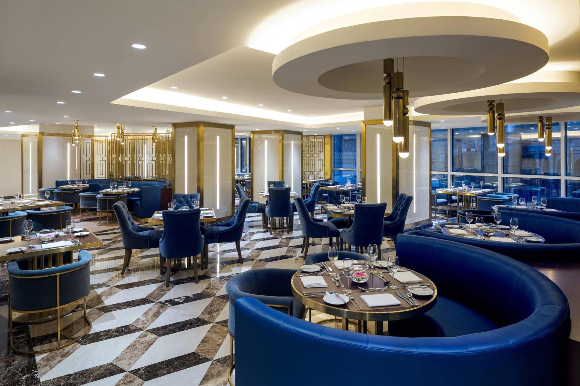 โรงแรมเชอราตัน มักกะฮ์ จาบาล อัล กาบา Mecca ภายนอก รูปภาพ The photo shows the interior of a modern restaurant. The design features elegant curved blue seating arranged around round tables with plates and glassware set for dining. The flooring is a striking marble pattern, adding to the upscale ambiance. The